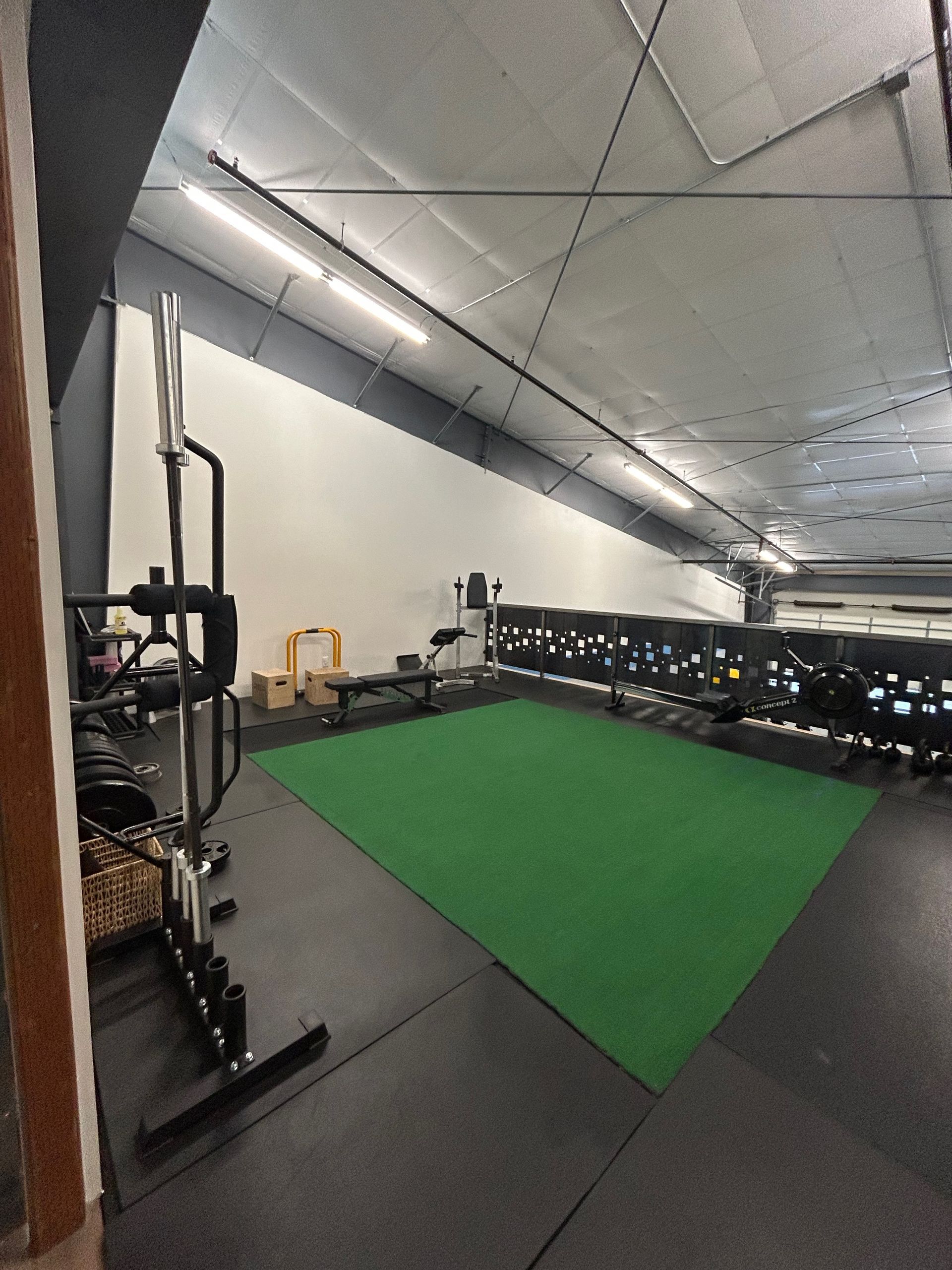 A home gym featuring a green turf section, black rubber flooring, and a squat rack with barbells under overhead lighting.