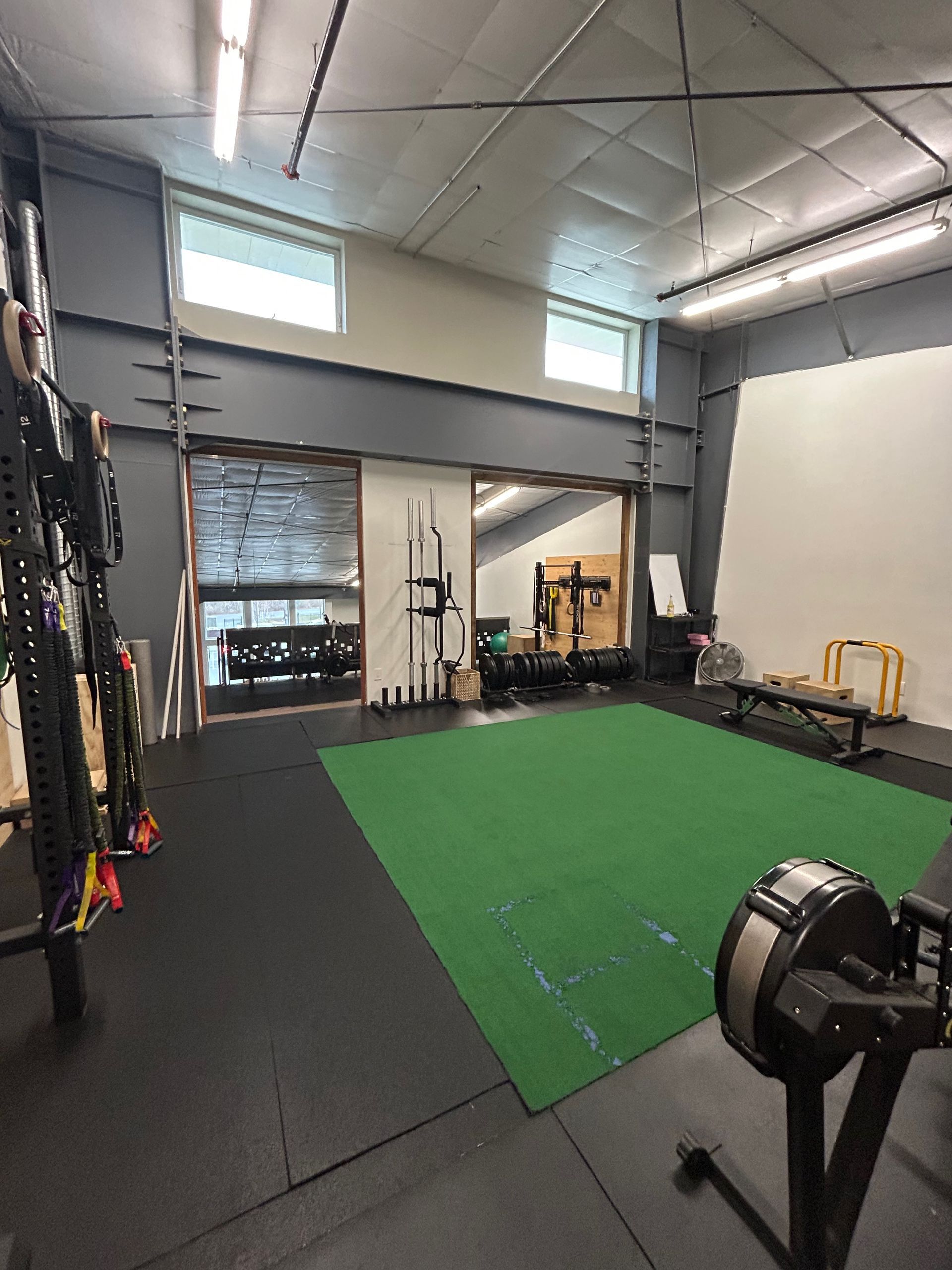 Interior of a gym with a green turf area, weight racks, barbells, a rowing machine, and a bench on black rubber flooring.