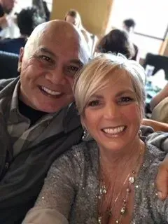 Two smiling people posing for a selfie indoors, with a softly lit background.