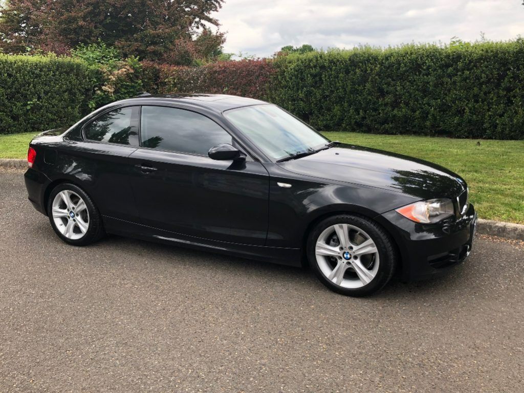Black sedan parked on a paved road beside a hedge on a cloudy day
