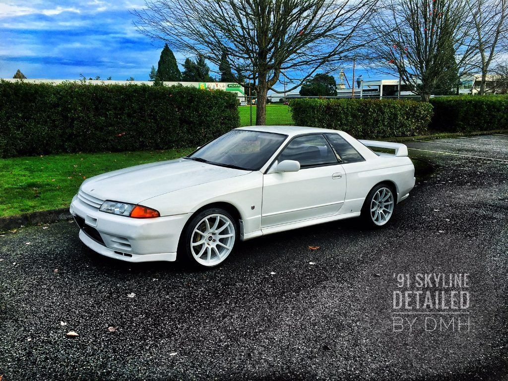 White Nissan Skyline coupe parked on wet asphalt beside a hedge under a cloudy blue sky
