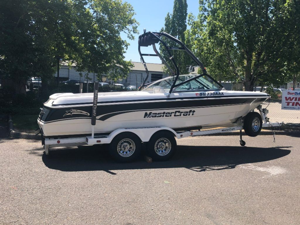 White MasterCraft boat on a trailer in a parking lot, with a wakeboard tower and trees in the background