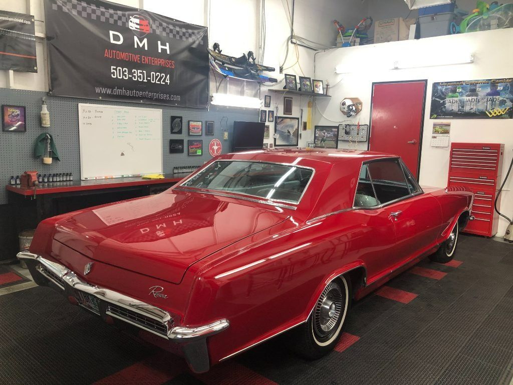 Red classic car parked in a garage with tools, shelves, and a red tool chest nearby
