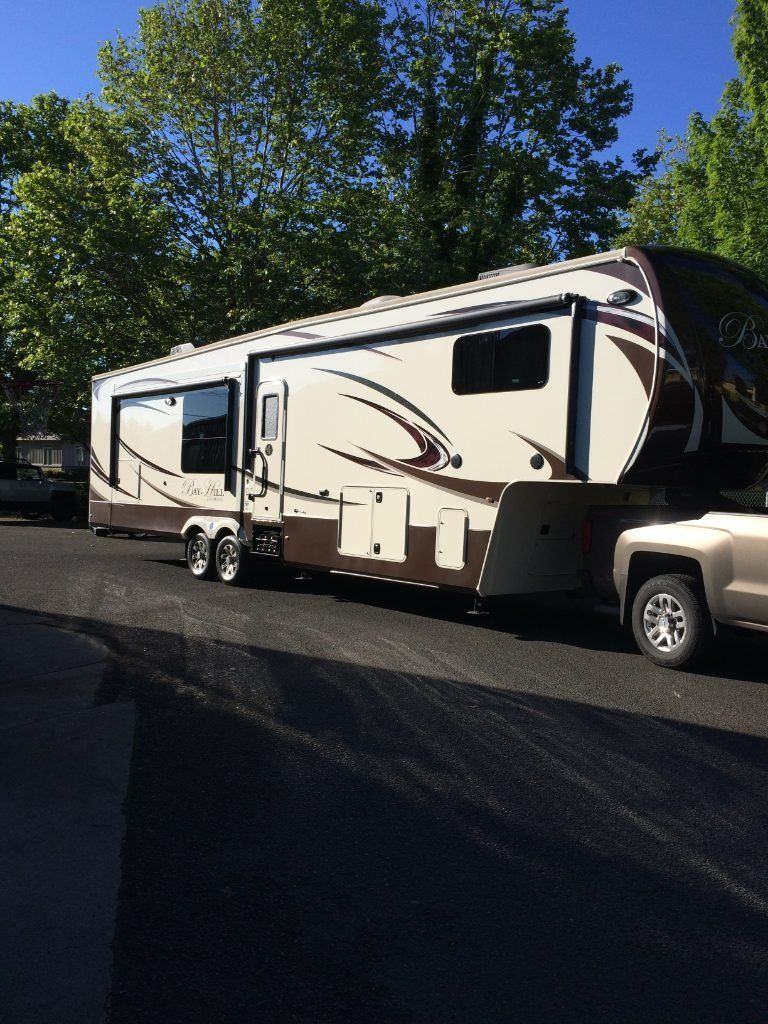 Beige and brown fifth-wheel RV parked on a paved lot beside trees in daylight