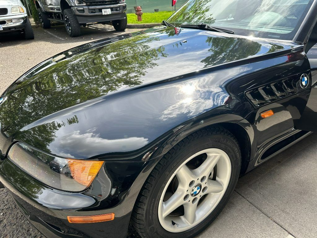 Black sports coupe parked outdoors, front-left view, with silver rims and reflected trees on the hood.