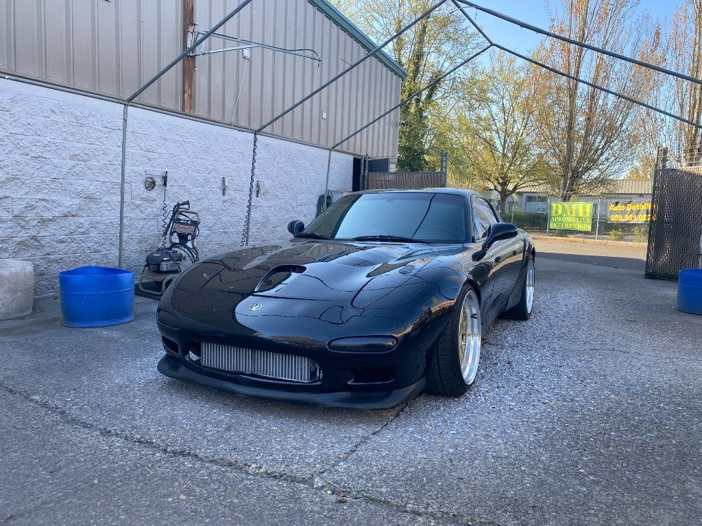 Black sports car parked on gravel beside a building, with a blue barrel and trees in the background