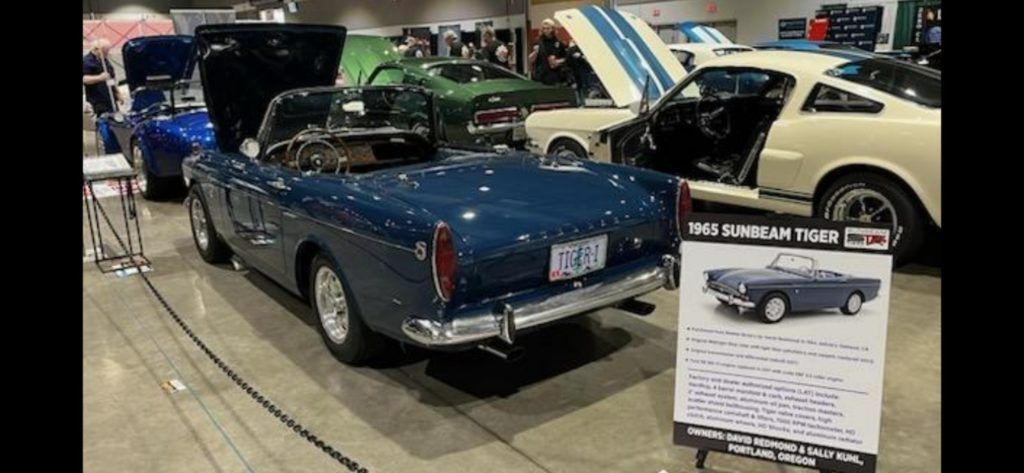 Blue classic convertible car displayed at an indoor auto show with a spec sign beside it
