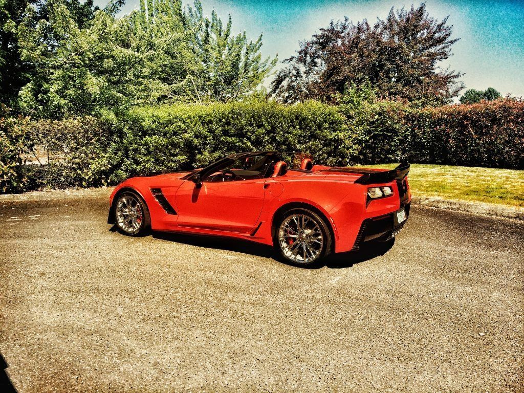 Red convertible sports car parked on a paved lot with trees and bushes in the background.