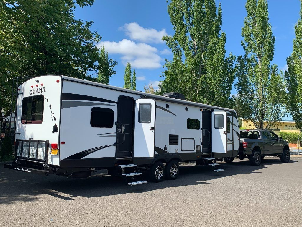 White RV with black and gray accents parked in a lot beside a pickup truck under tall trees.