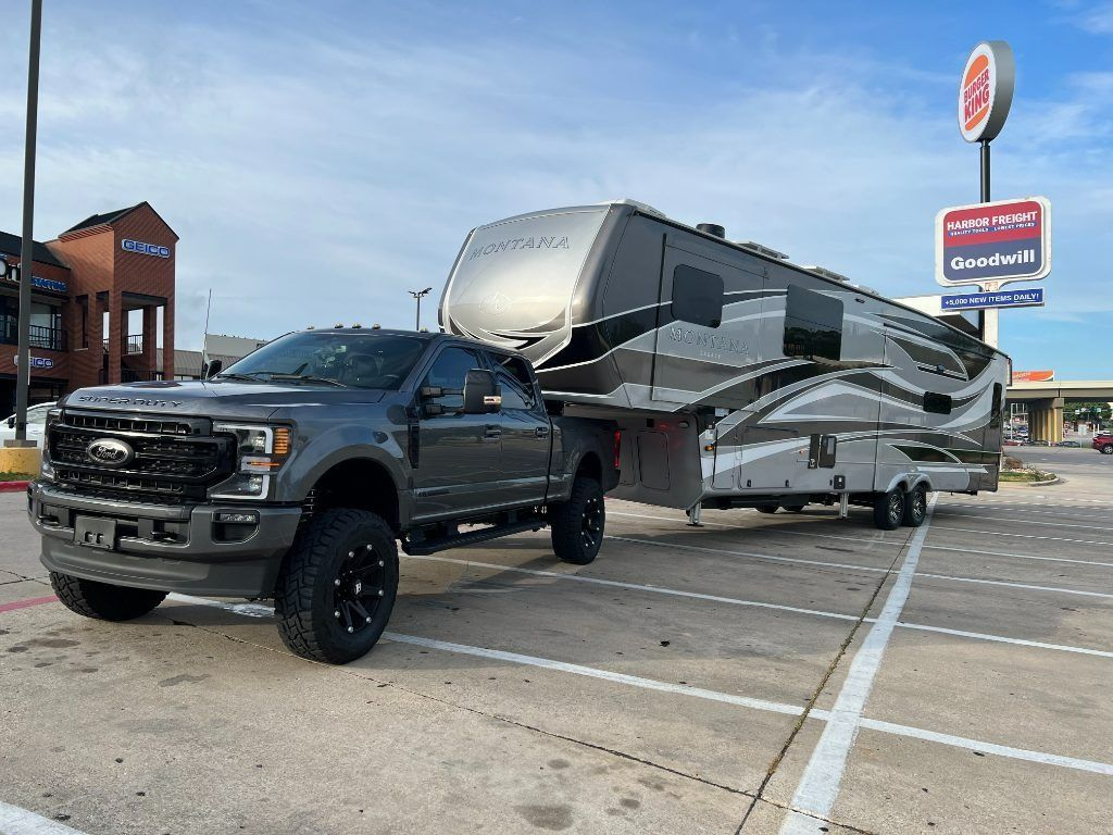 Black pickup truck towing a large gray fifth-wheel RV in a parking lot near a roadside sign.