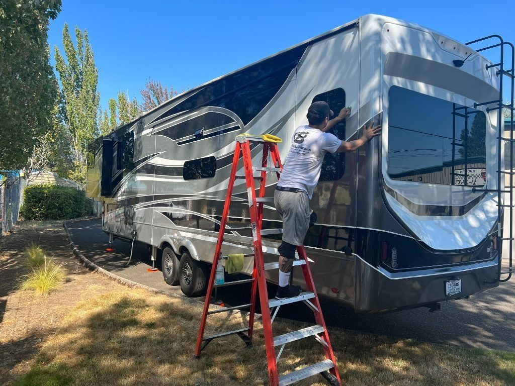 Person on a ladder washing a silver and black RV outdoors in a sunny yard