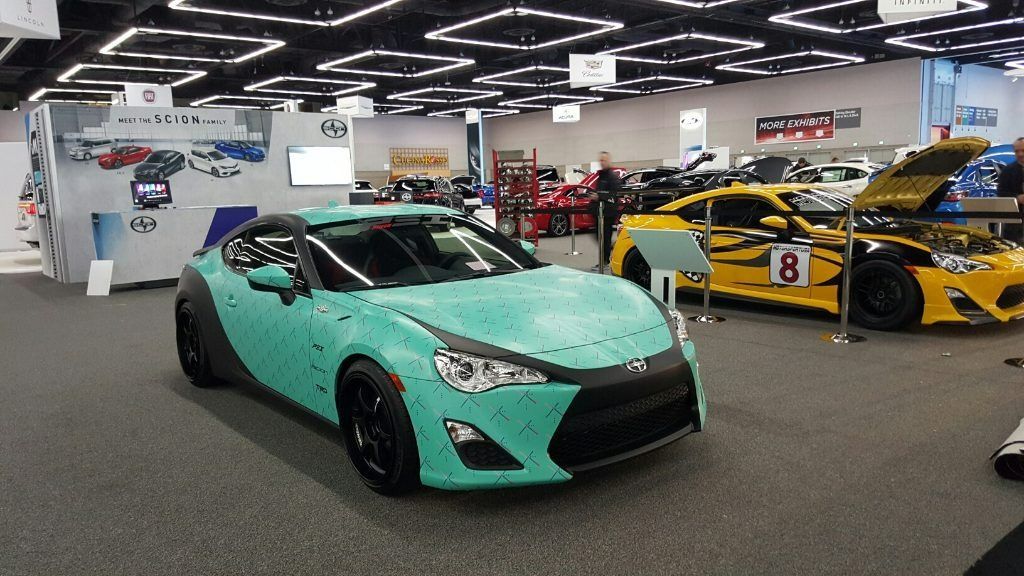 Mint green sports car in an indoor showroom, with yellow race cars in the background.