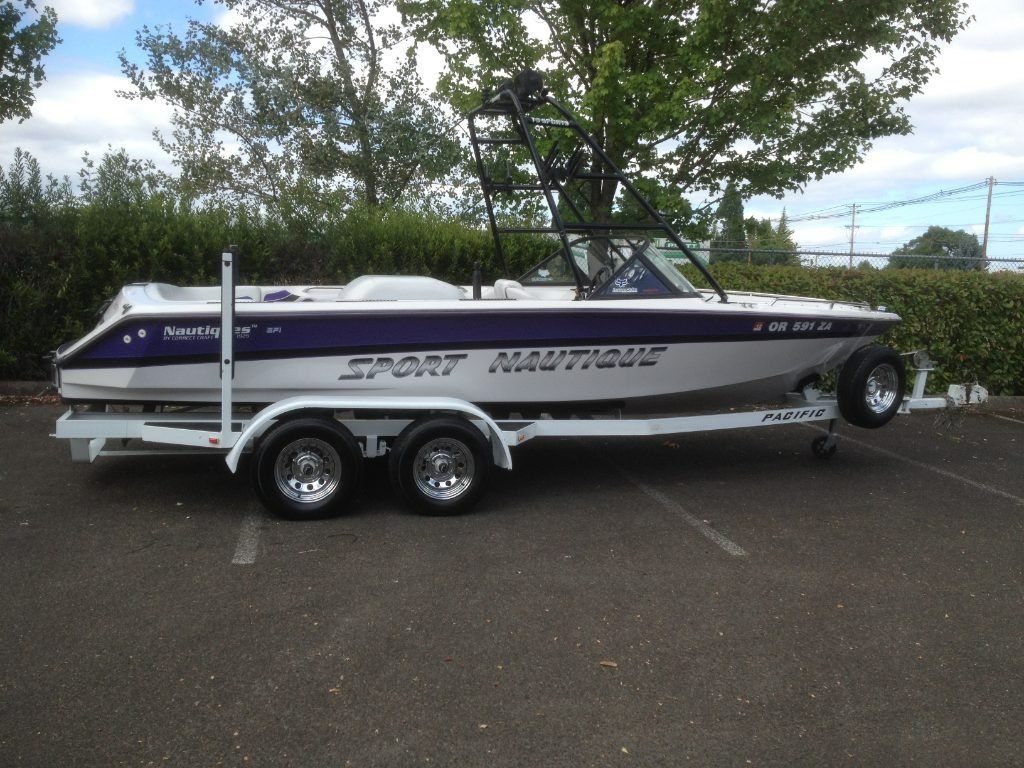 White and purple Speedster boat on a trailer in a parking lot with trees and hedges behind it