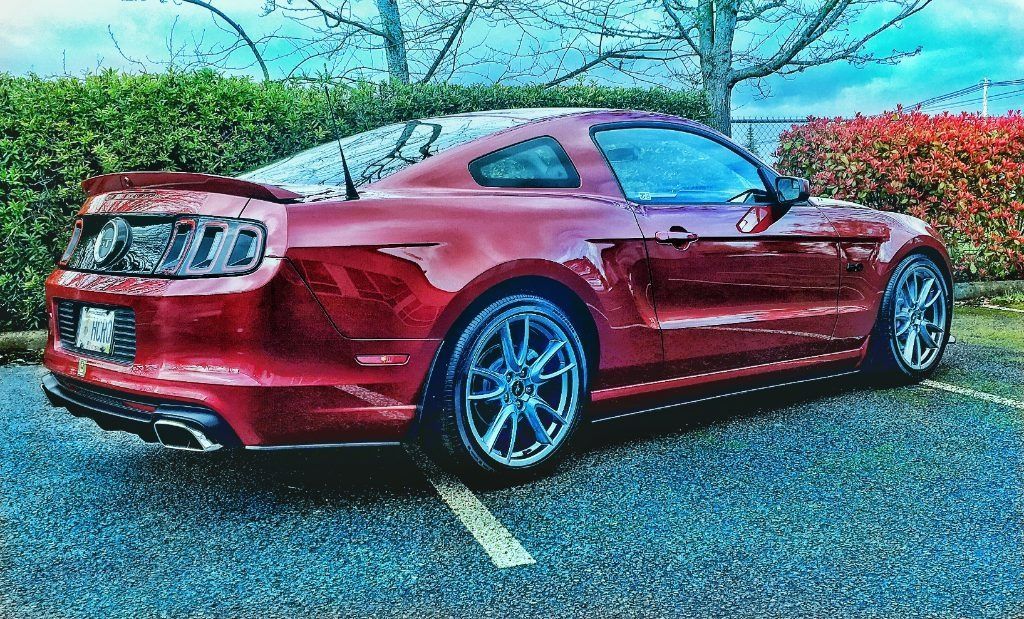Red sports coupe parked in a lot, with blue rims and trees and shrubs in the background.