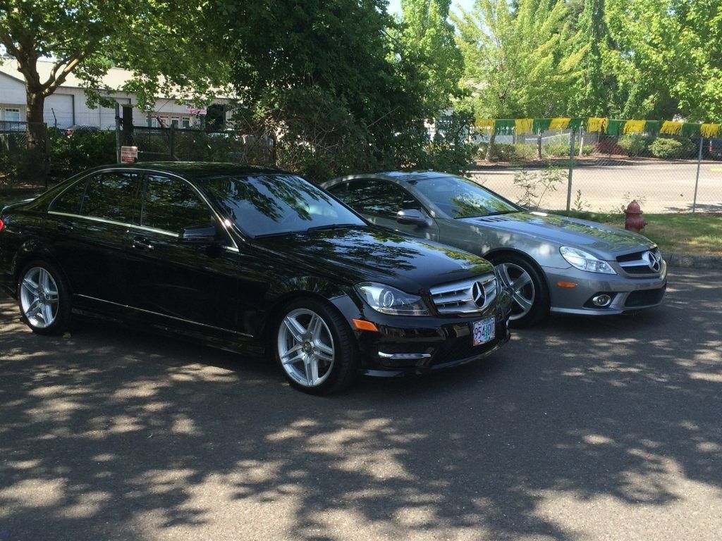 Two parked cars, a black sedan and a silver sedan, on a shaded street near trees and a fence