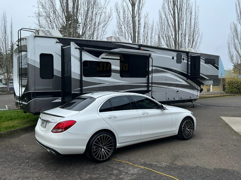 White sedan parked beside a gray-and-black RV in an outdoor lot, with tall trees in the background