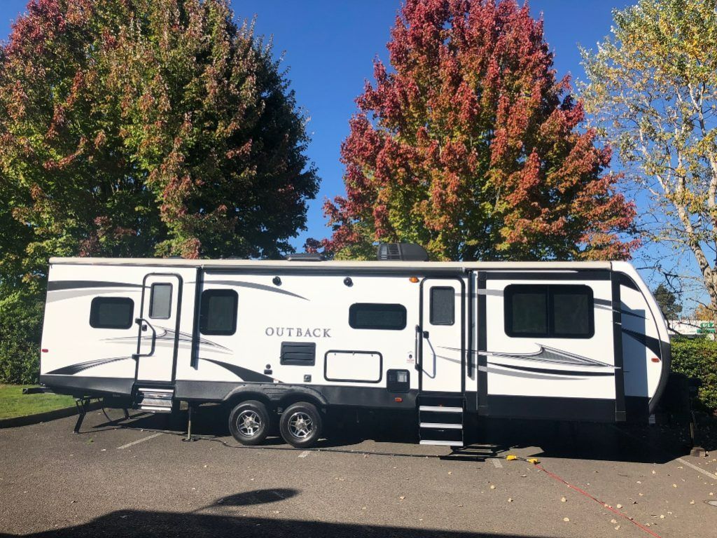 White and black travel trailer parked in a lot under autumn trees with red and green leaves