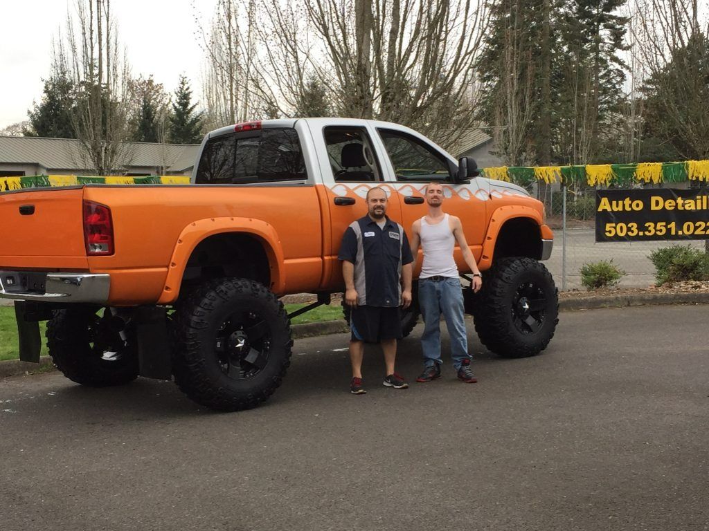 Two people standing beside a lifted orange pickup truck in a parking lot