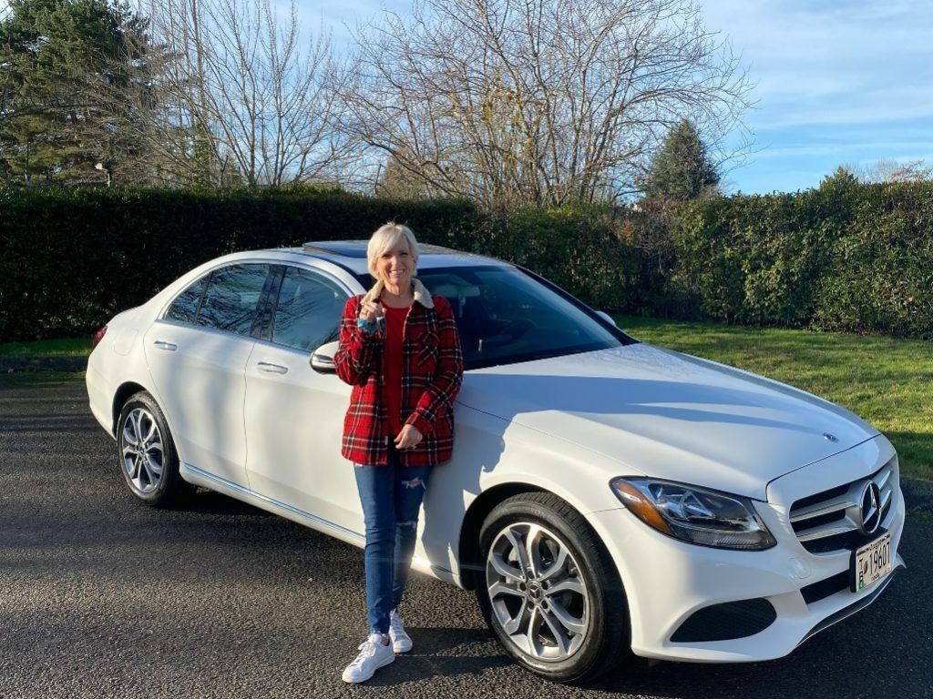 Person standing beside a white sedan on a road, holding a red phone, with hedges and trees in the background