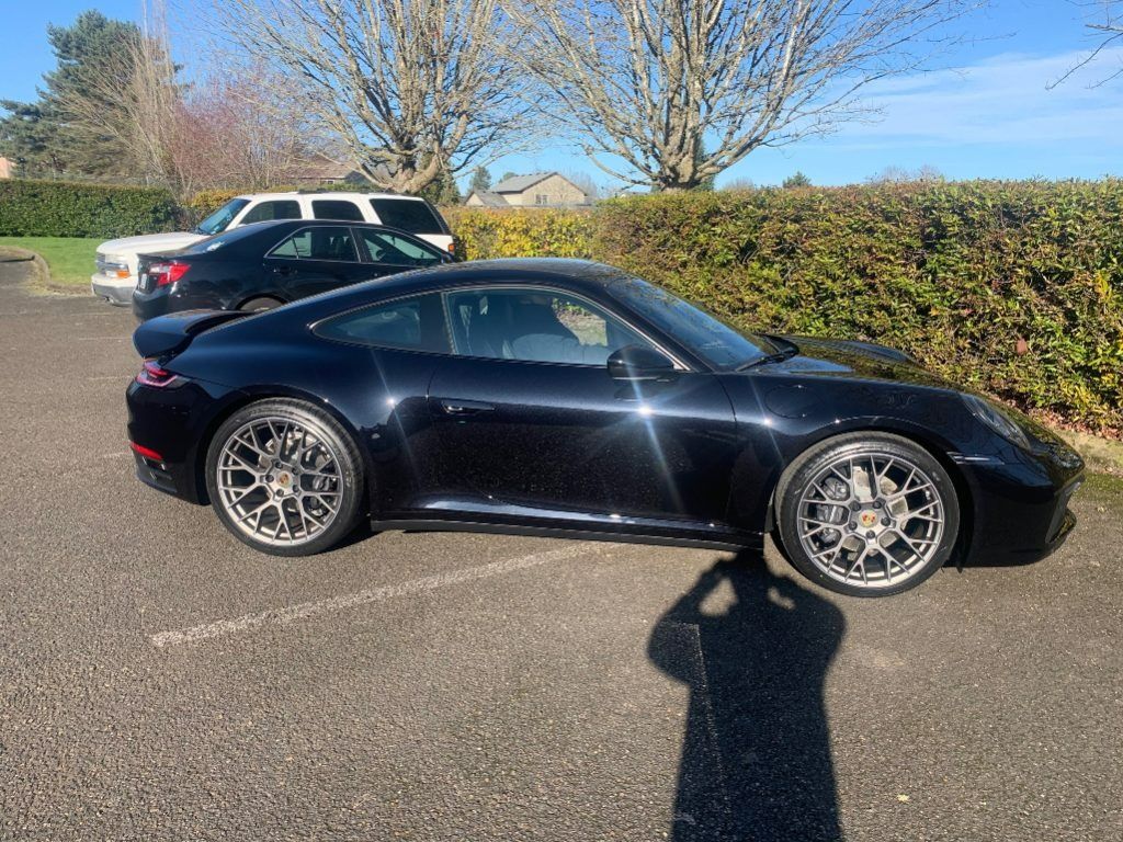 Black sports coupe parked outdoors in a lot, with a hedge and blue sky in the background.