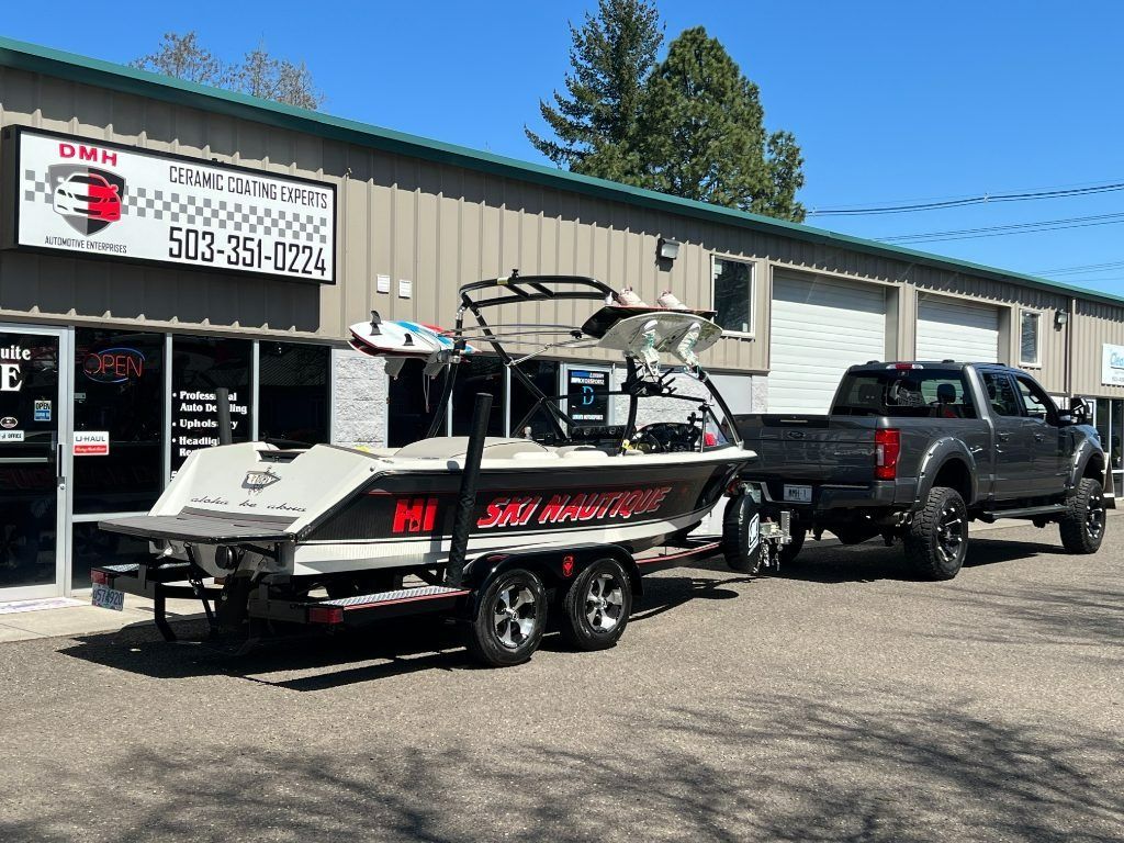 Black wakeboard boat on a trailer hitched to a dark pickup outside a building