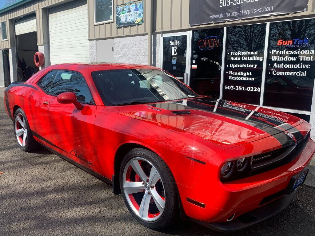 Red Dodge Challenger parked outside a shop, with black racing stripes and red rims.