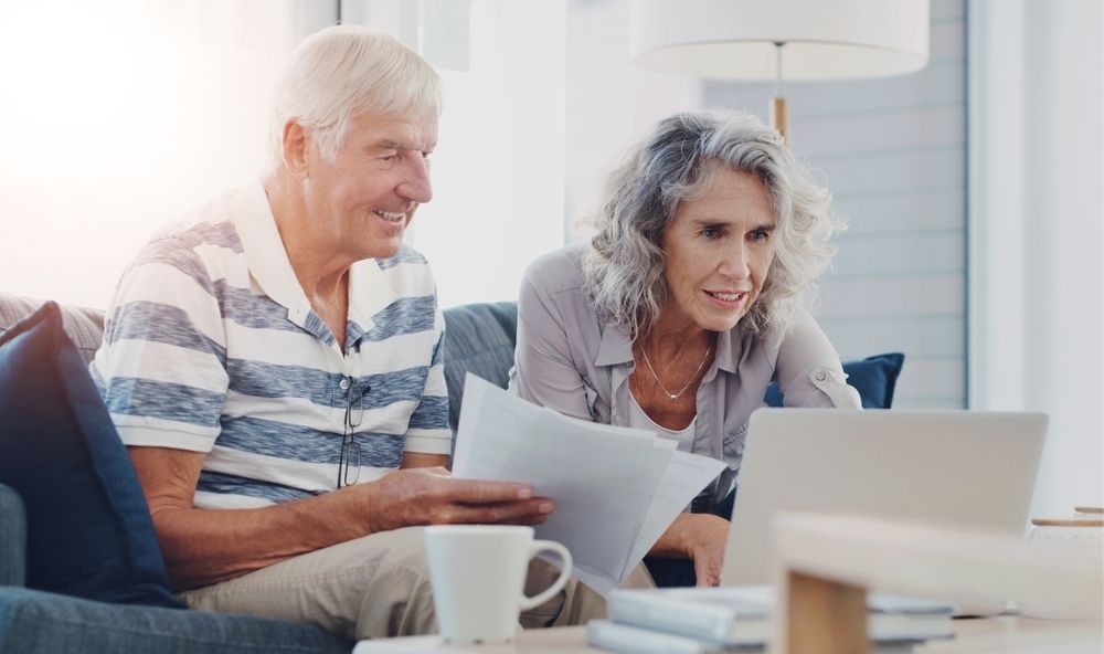 An elderly couple is sitting on a couch looking at papers and a laptop.
