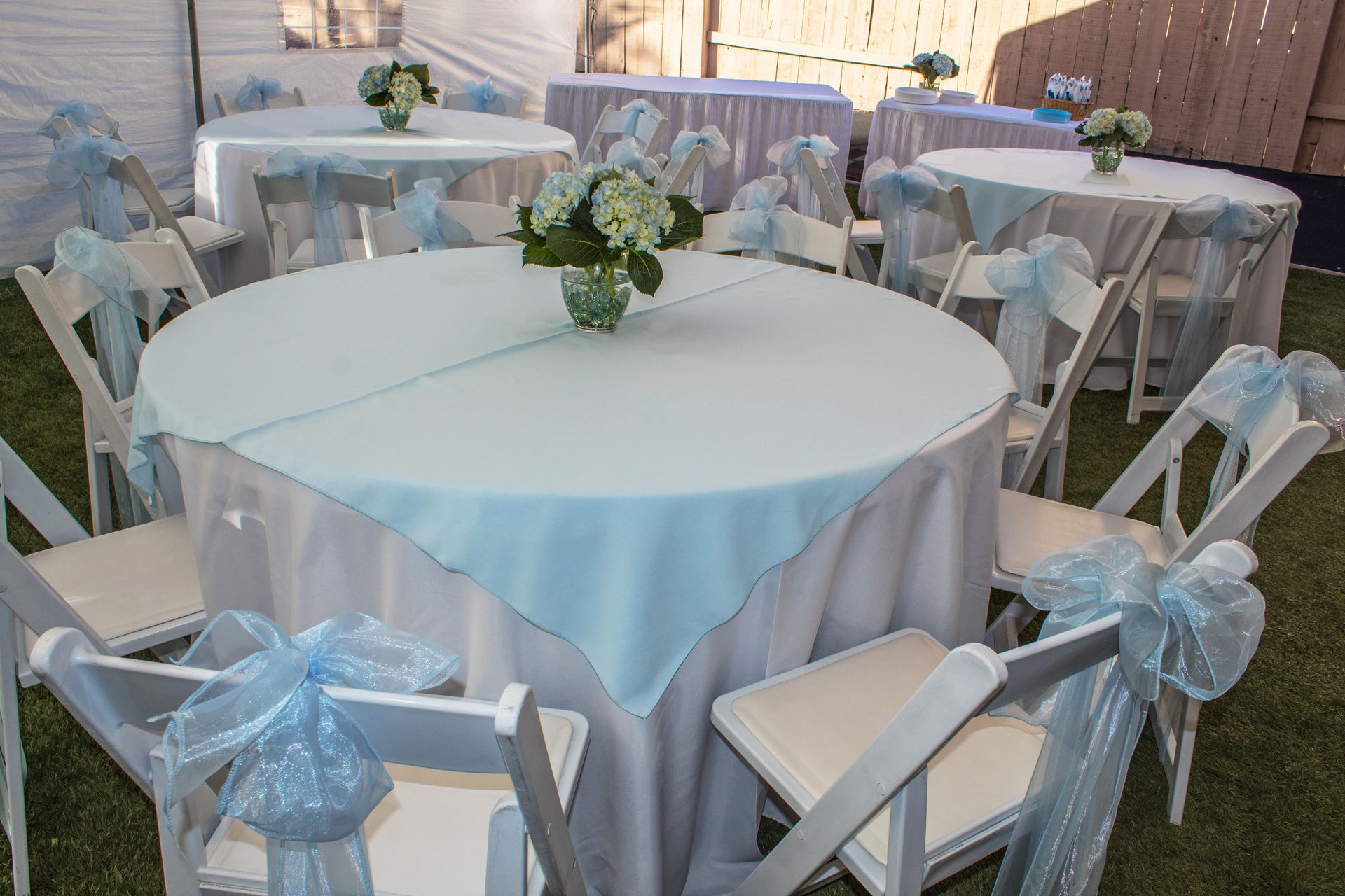 Round white-draped reception tables with blue bows and floral centerpieces outdoors