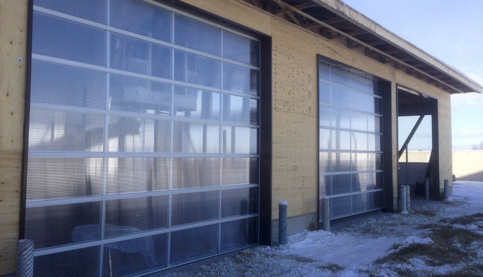 Three clear, paneled garage doors on a light-colored wooden building with snow on the ground.
