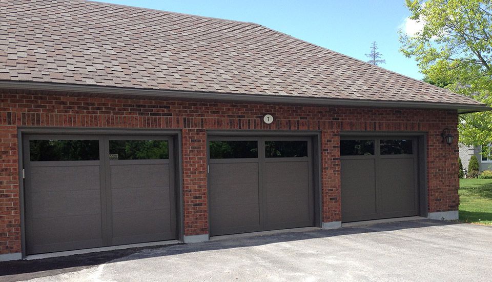 Three-car garage with brown doors, brick exterior, and a shingled roof under a clear, blue sky.
