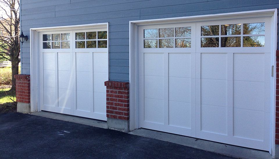 Two white garage doors with glass panel tops, on a blue-sided building with brick accents.