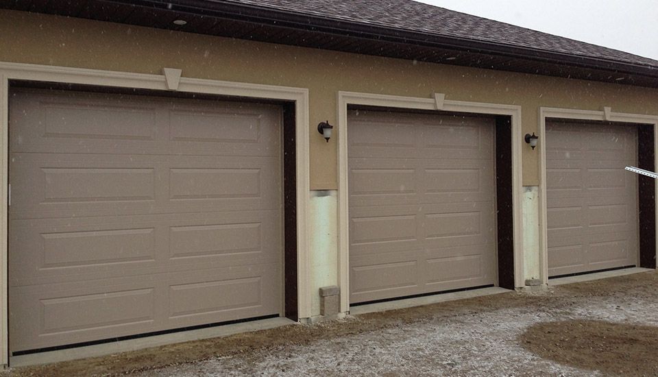 Three beige garage doors on a light brown building, snowy conditions.