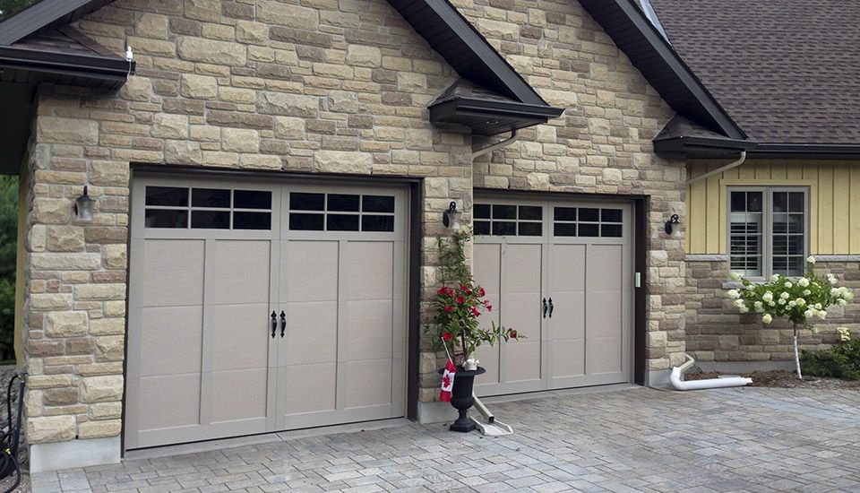 Two-car garage with stone facade and beige doors. Brick driveway with small flowers and shrubs.