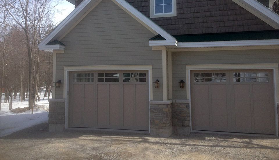 Two-car garage with tan doors, stone columns, and light green siding. Snow-covered ground and trees in background.