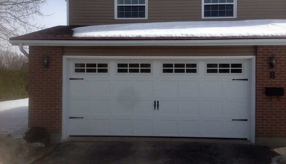 White garage door with windows, black hardware, set in a brick and siding facade. Snow on the roof.