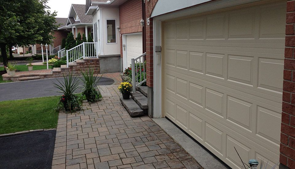Brick walkway leading to houses with garage and steps. Beige garage door on right. Green grass, trees.