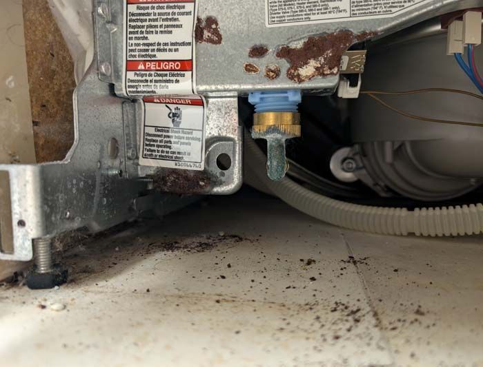 Dishwasher's water inlet valve, corroded and dirty. Metal frame with warning labels, visible plumbing.