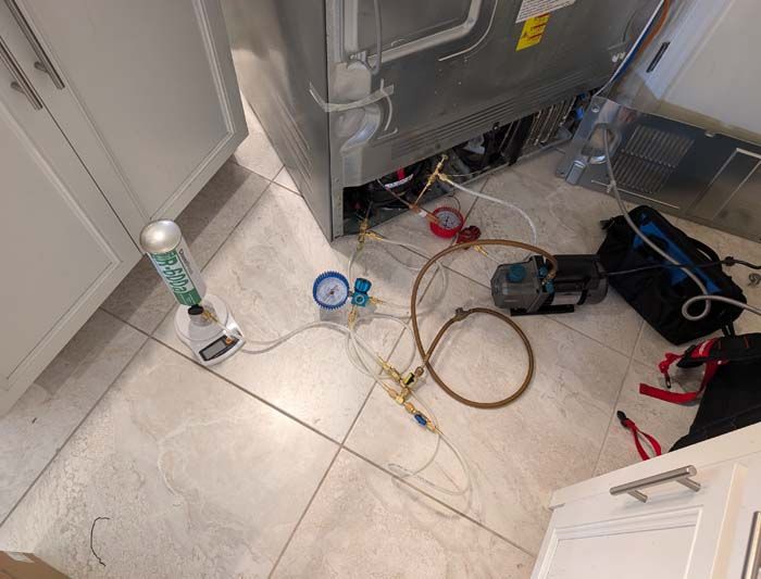 Refrigeration repair setup: gauges, vacuum pump, and refrigerant canister near a refrigerator in a kitchen.