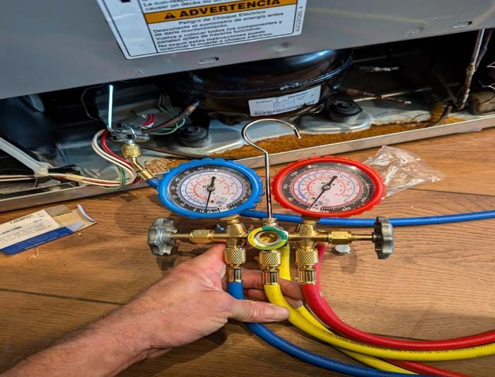 Hand holding gauges connected to a refrigerator, checking pressure. Blue and red hoses, and two gauges.