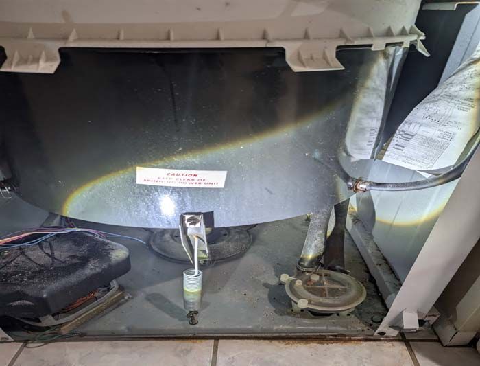 Interior view of a washing machine's base, showing mechanical components. Silver, white, and grey parts are visible.