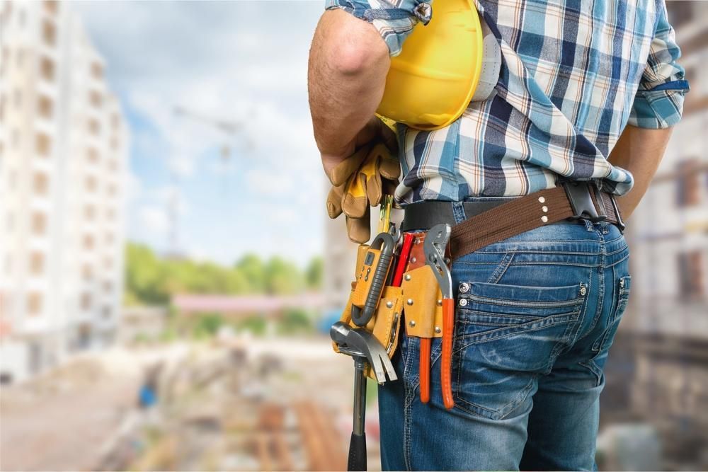 Construction Worker In Blue Jeans And Plaid Shirt, Holding Yellow Helmet And Tools At A Construction Site