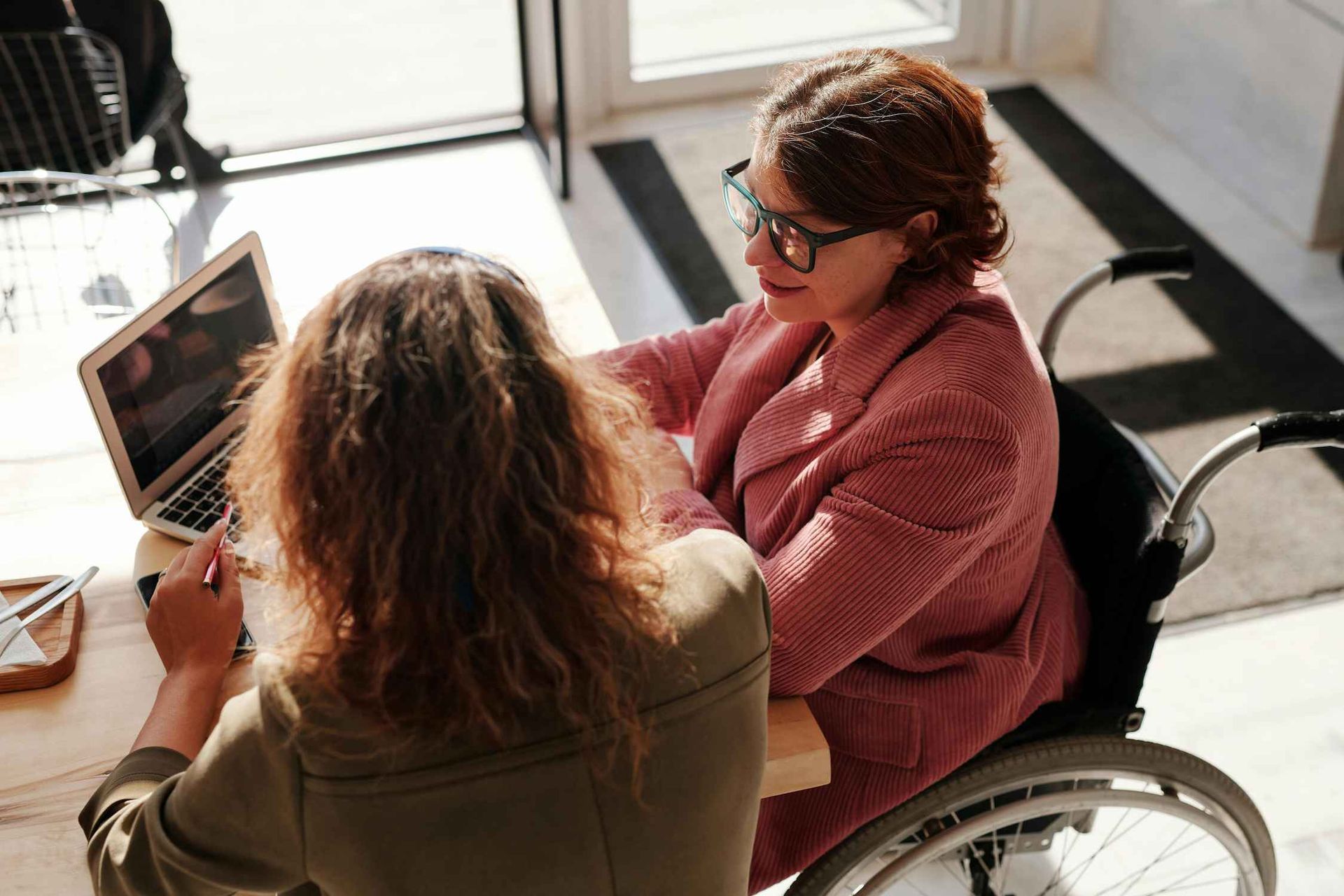 a Woman in a Wheelchair is Sitting at a Table With Another Woman Using a Laptop Computer — Coast & Country Insurance Consultants Pty Ltd In Central Coast, NSW