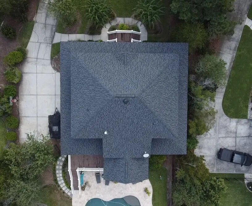 Overhead view of a house with a dark gray roof, driveway, trees, a pool, and a black car. Overhead view of a house with a dark gray roof, driveway, trees, a pool, and a black car.