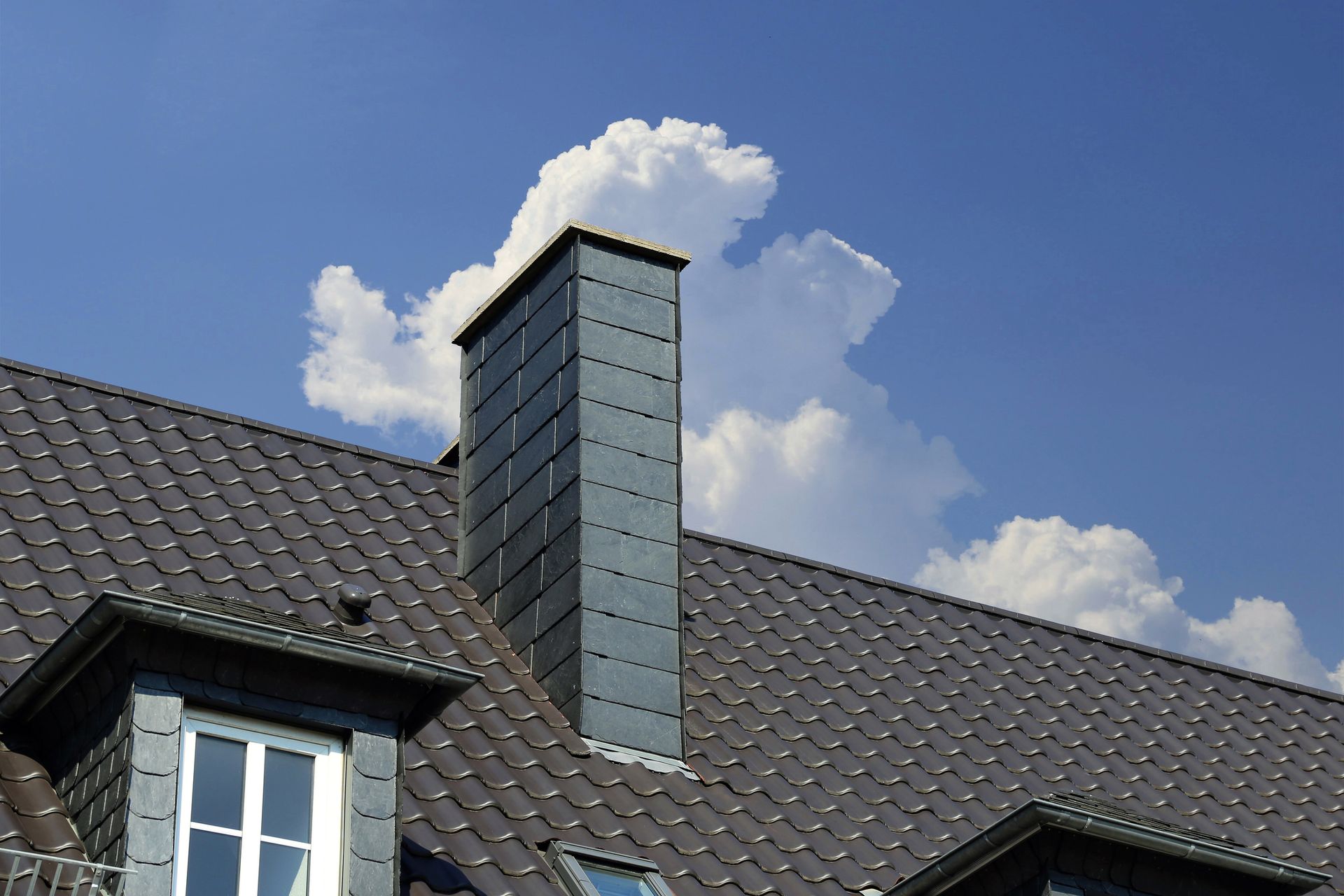 Dark gray roof with a tall chimney against a blue sky with fluffy white clouds.