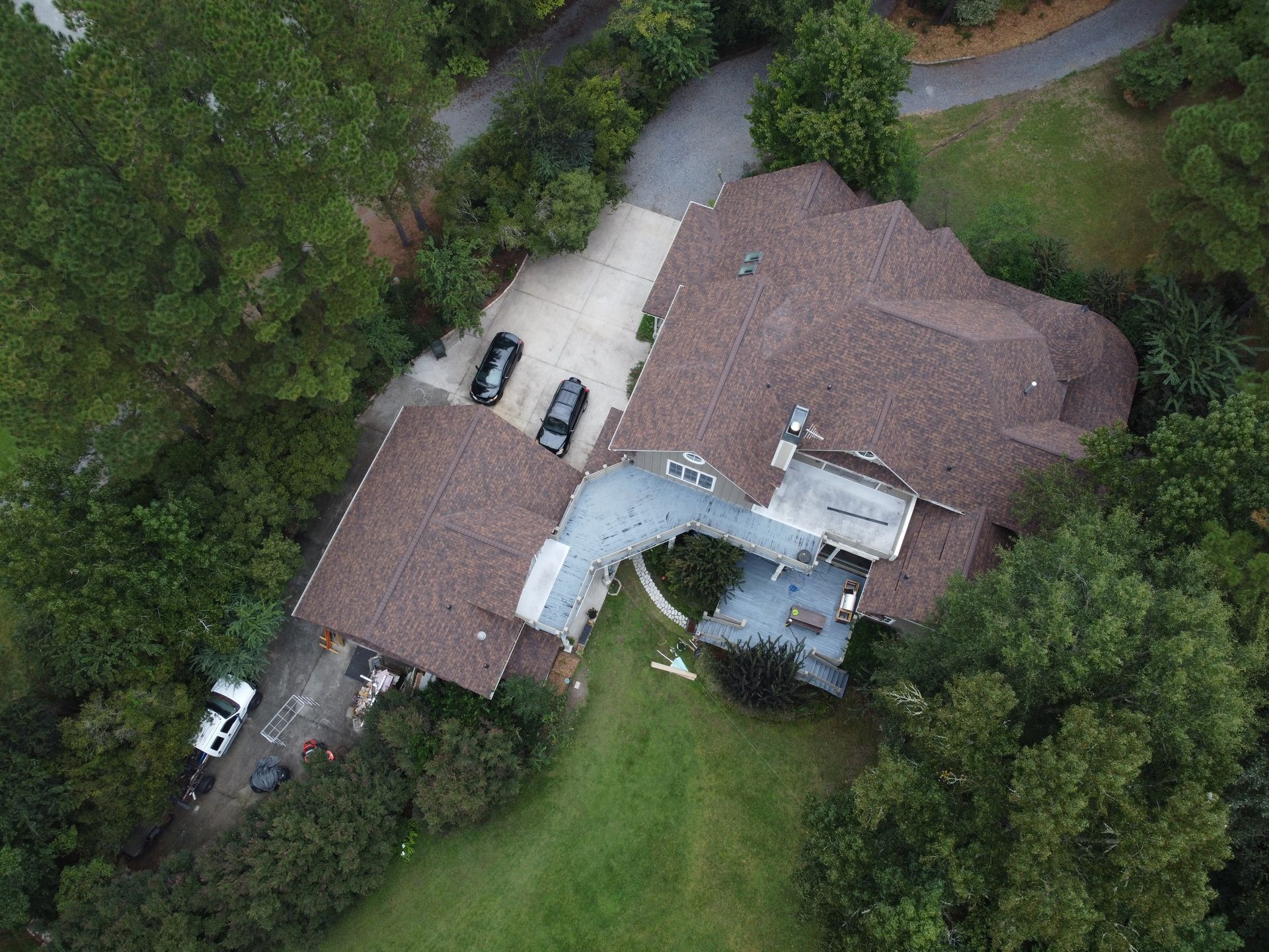 Aerial view of a brown-roofed house with a driveway, surrounded by trees and a green lawn.