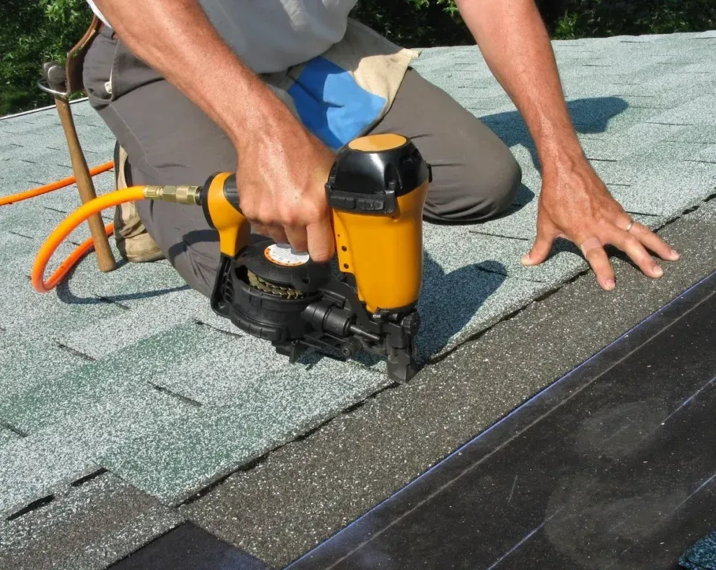 Person kneeling on a roof, using a nail gun to install roofing shingles.