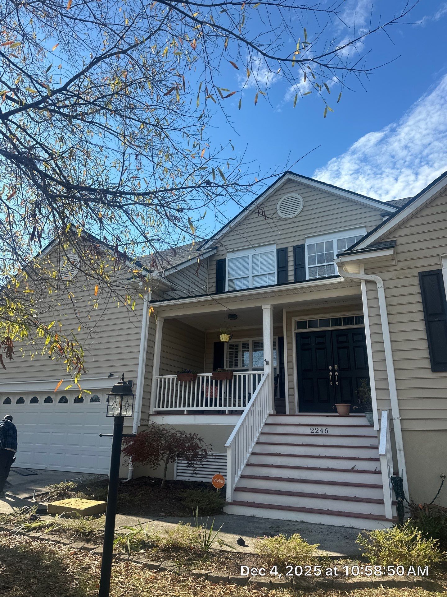 Beige house with a porch and steps leading to the front door, tree branches overhead, and a blue sky.