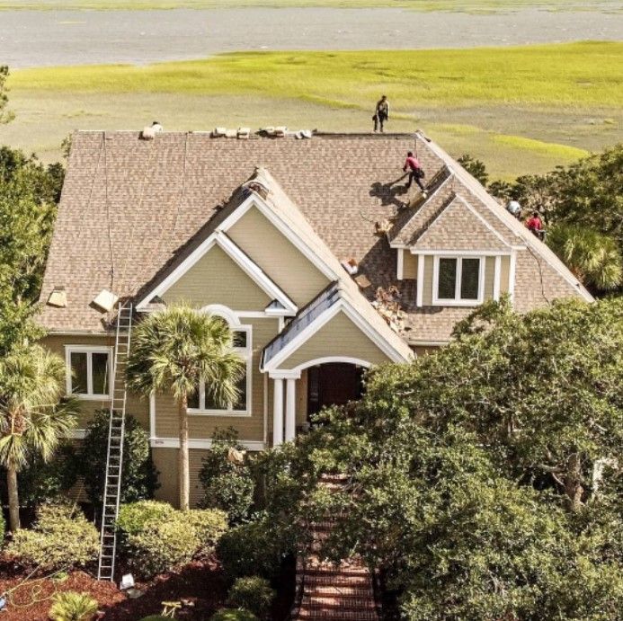 House with workers on the roof, trees in foreground. Ladder on side of house, green and brown tones.