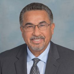 Portrait of a smiling man in a dark suit, light blue shirt, and patterned tie against a gray-blue backdrop