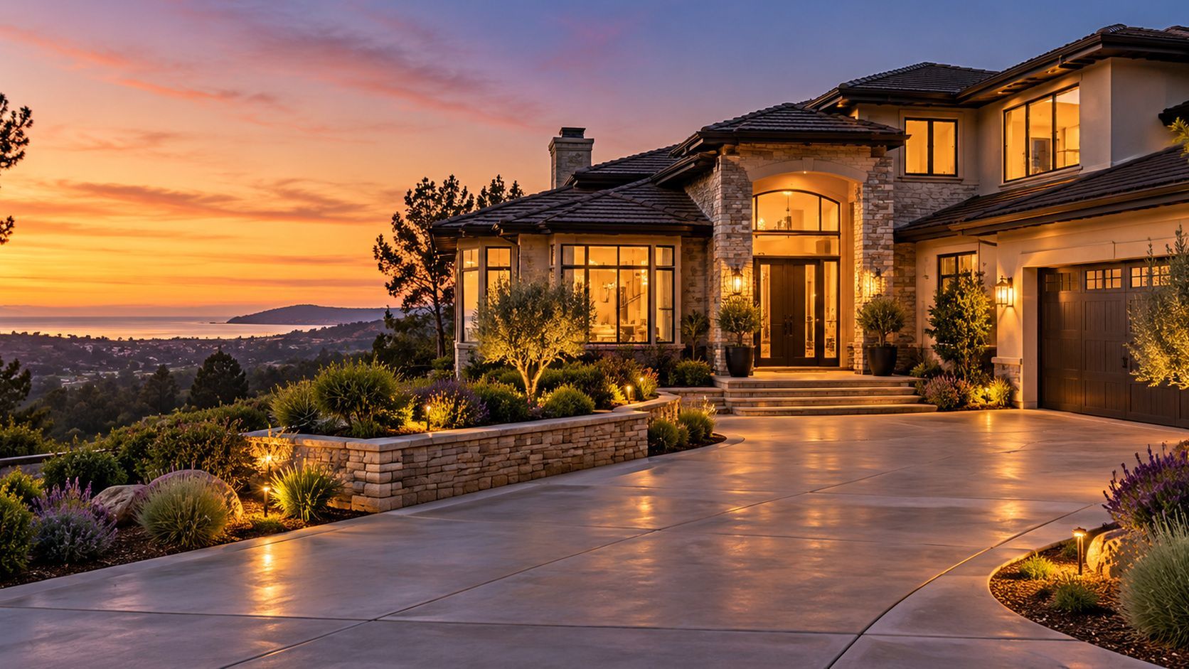 Luxury hillside home at sunset with warm lights, stone facade, and a winding driveway overlooking a scenic valley
